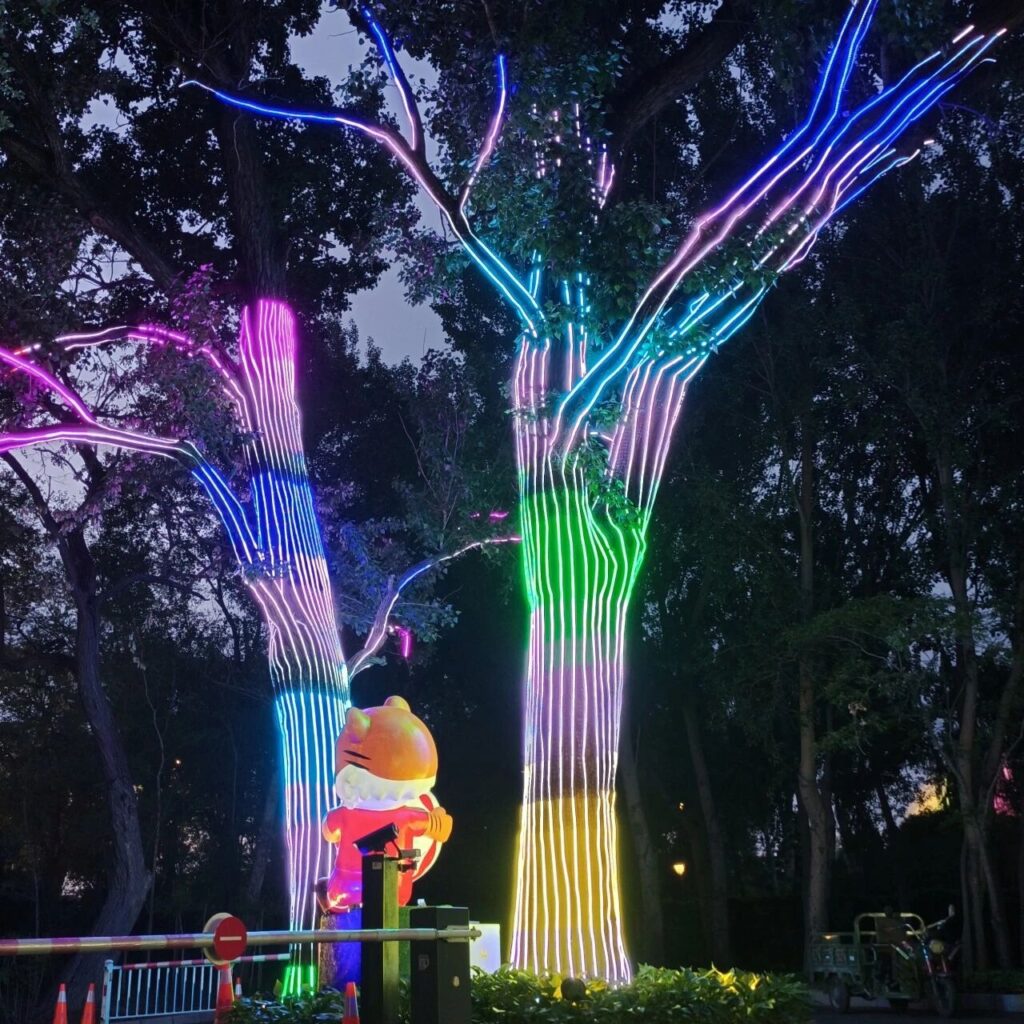 Duas árvores cênicas com tronco e galhos revestidos por LED RGB em degradê de cores, formando um marco visual noturno em parque