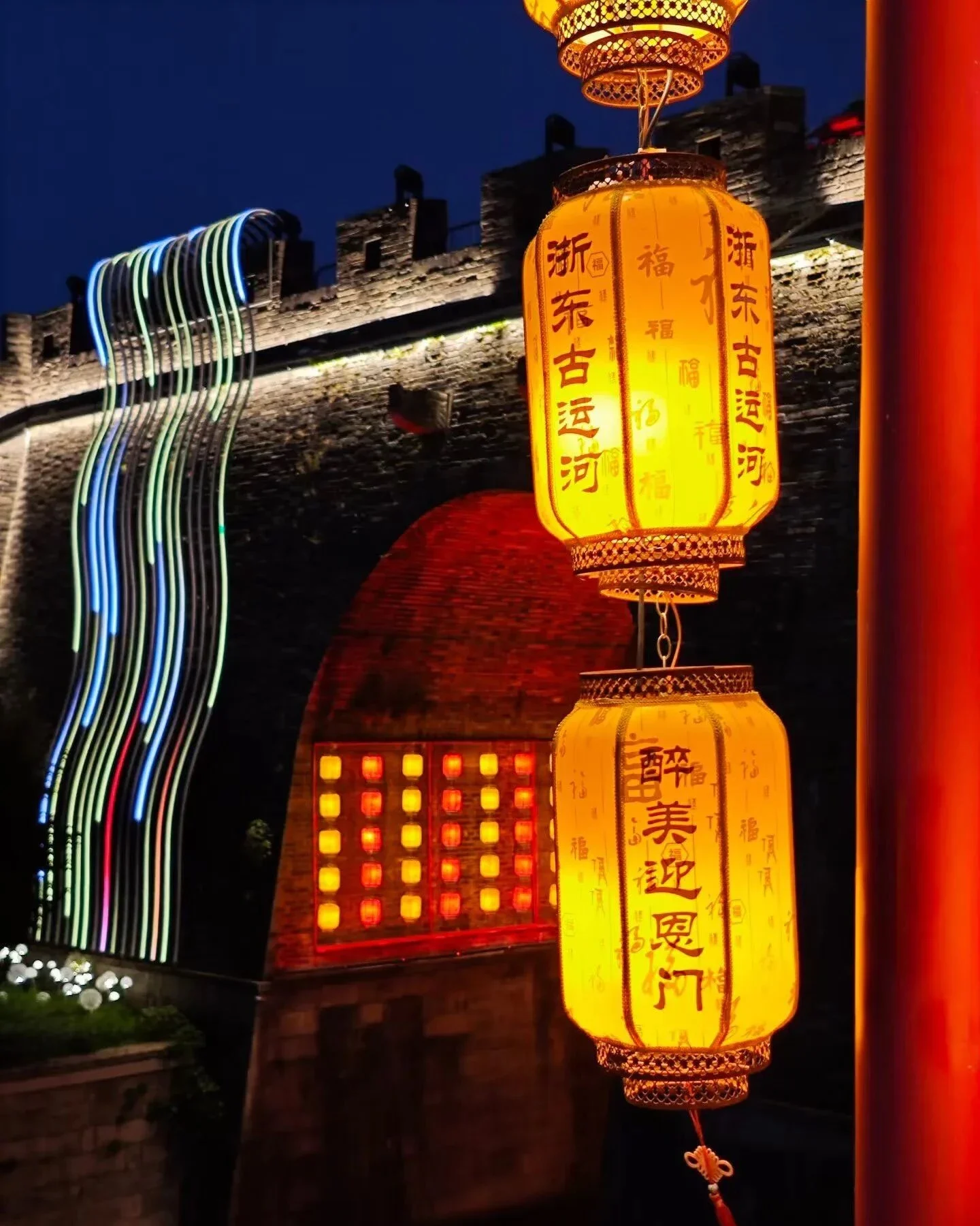 Duas grandes lanternas chinesas amarelas iluminadas em frente a um portão de muralha histórica, com rastros de luz coloridos ao fundo