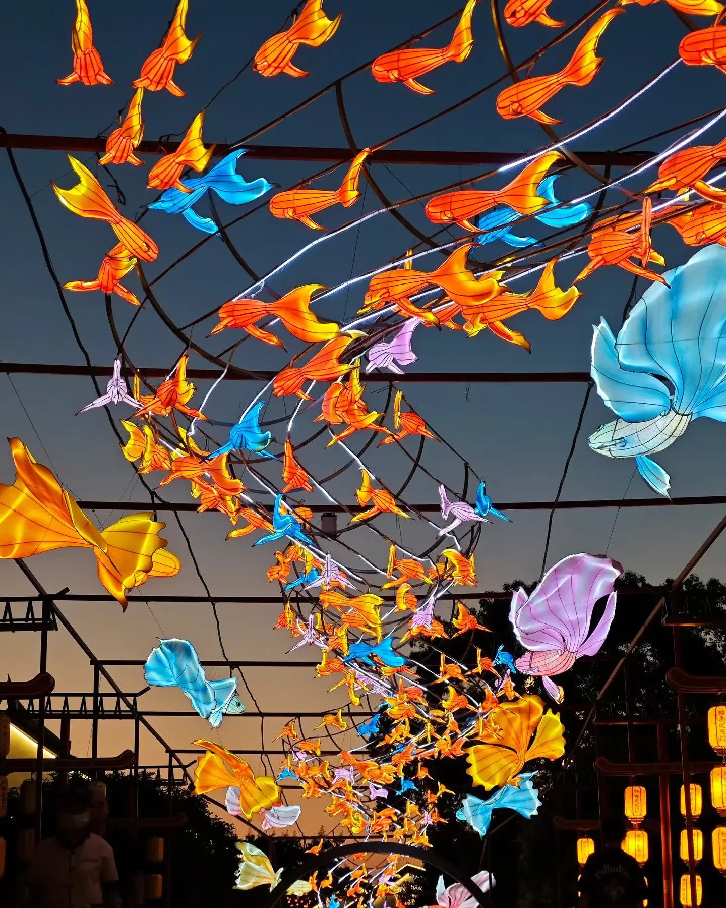 Cobertura de rua tomada por lanternas em forma de peixes alaranjados e borboletas azuis iluminadas, sobre um mercado noturno ao entardecer